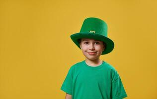 peu garçon portant vert T-shirt et lutin irlandais casquette regards à le caméra. Saint patrick journée. Jaune Contexte avec copie espace photo