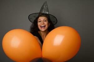 Latin américain femme habillé dans noir et sorcière chapeau coups d'oeil par le Orange gonflé des ballons et sourit avec une magnifique à pleines dents sourire à la recherche à le caméra. Halloween concept avec copie espace photo