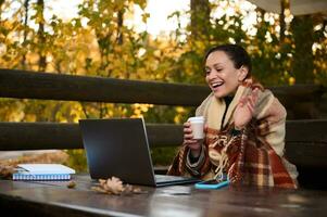 de bonne humeur âge moyen femme chauffage se enveloppé dans plaid couverture et en buvant chaud café de papier tasse pendant vidéo appel tandis que séance sur ouvert pays terrasse sur cool l'automne journée dans un chêne bosquet photo