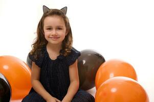 de bonne humeur séduisant adorable peu fille dans noir carnaval robe et cerceau avec chat oreilles séance sur une blanc surface avec bi coloré des ballons, sourit à la recherche à caméra. Halloween décoration. copie espace photo