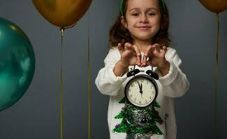 doux concentrer sur minuit sur le l'horloge visage sur un alarme dans le mains de magnifique de bonne humeur fille souriant joliment à la recherche à caméra contre gris Contexte avec copie espace pour Noël publicité photo