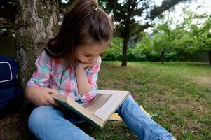 adorable diligent peu enfant fille, premier classe étudiant lit une livre dans le parc, séance en dessous de le arbre sur herbe. photo