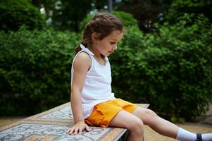 caucasien content peu enfant fille 4 ans vieux, dans été porter, pièces et sourit séance sur mosaïque banc dans le ville parc photo