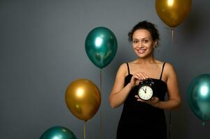 élégant mixte course femme habillé dans soir robe, détient un alarme l'horloge et mignonne sourit avec à pleines dents sourire à la recherche à caméra, contre air des ballons sur gris Contexte. c'est minuit, joyeux Noël photo