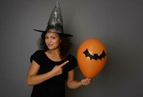 mignonne souriant magnifique et joyeux femme habillé dans Halloween carnaval noir sorcière vêtements, points sur un Orange air Balle avec une Fait main chauve souris, permanent isolé plus de gris Contexte avec copie espace photo