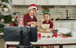 adorable peu des gamins blogueur cuisiniers, une garçon verse Lait dans une bol et une fille pétrit pâte, enregistrement une pas à pas vidéo de une recette pour sa vlogging sur une téléphone intelligent monté sur une trépied. photo