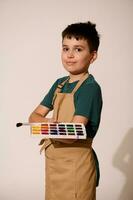 adolescent garçon dans beige tablier, détient pinceau et palette avec aquarelles, sourit à caméra, isolé studio Contexte photo