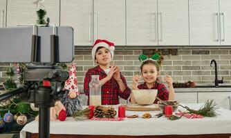 magnifique les enfants cuisiniers, mignonne fille et Beau garçon portant Père Noël chapeau à la recherche à caméra dans téléphone intelligent monté sur trépied, enregistrement vidéo pour vlogging, cuisine Noël pain ensemble dans cuisine photo