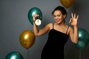 de bonne humeur mixte course femme détient alarme horloge, c'est minuit, montrant D'accord signe souriant à la recherche à caméra, isolé sur gris Contexte avec des ballons. Nouveau année, joyeux Noël concept pour La publicité photo