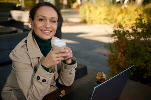 sur de soi portrait de une charmant affaires femme en portant une à emporter artisanat papier tasse avec chaud boisson et souriant à pleines dents sourire à la recherche à caméra séance sur en bois banc de Extérieur café dans l'automne chêne bosquet photo