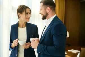 deux affaires les partenaires, attrayant Jeune femme et charmant homme dans affaires combinaisons, Regardez à chaque, en portant tasse de café, permanent par une fenêtre dans Hôtel chambre. concept de l'amour affaires et intimité à travail photo