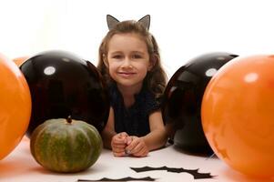 souriant adorable enfant fille dans carnaval costume, peu sorcière mensonge vers le bas sur blanc Contexte avec copie espace suivant à citrouilles, Fait main chauves-souris et coloré Orange noir des ballons. Halloween fête concept photo