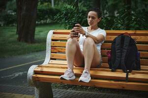 Jeune ethnique femme chèques mobile app sur sa téléphone intelligent, défilement nouvelles alimentation, séance sur une en bois banc dans le ville parc photo