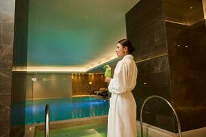 milieu vieilli jolie femme dans blanc éponge peignoir de bain permanent par le bassin et jacuzzi dans luxe santé spa, avec une fraîchement pressé jus dans sa mains. bien-être, spa, des loisirs, relaxation concept photo