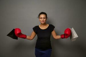 magnifique souriant africain femme portant rouge boxe gants détient achats Sacs, joliment sourit à la recherche à caméra contre gris mur Contexte avec copie espace pour noir Vendredi publicité. coup à des prix concept photo