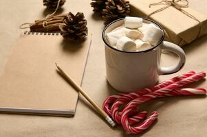 Noël composition avec cadeau dans artisanat emballage papier, cannelle lié avec corde, pin cônes, en bois crayon sur bloc-notes, rayé sucettes bonbons cannes mensonge vers le bas près une tasse avec cacao et guimauves. photo