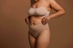 tondu vue de une femme dans beige sous-vêtements pose sa mains sur sa estomac avec détendu muscles cette avoir perdu leur Ton et étendue Des marques après grossesse et accouchement. corps positif concept photo