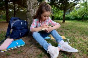 caucasien charmant peu enfant fille, une premier niveleuse l'écriture sur cahier d'exercices, engagé sur Faire devoirs après classe en plein air. photo