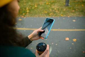 concentrer sur mobile téléphone avec noir vide Vide écran avec copie espace pour publicité dans le mains de flou mature femme en portant à emporter papier tasse et profiter l'automne temps repos dans forêt parc photo