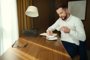 réussi fatigué caucasien homme d'affaire est assis à une bureau avec une tasse de délicieux fraîchement brassée café et écrit une affaires voyage rapport sur carnet de notes, des plans réunions, relaxant dans une Hôtel pièce après travail. photo