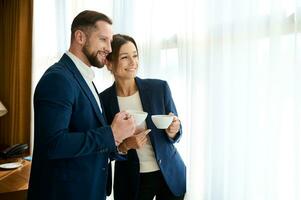 deux souriant charmant réussi affaires personnes, une Beau confiant homme et une magnifique femme dans affaires combinaisons, en portant une tasse de café et en toute confiance à la recherche en dehors le fenêtre à un côté. photo