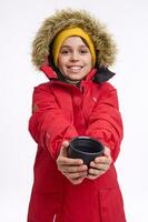 charmant de bonne humeur âge scolaire garçon dans brillant chaud vêtements sourit à pleines dents sourire, à la recherche à le caméra, chauffage le sien mains sur une tasse avec une chaud thé boire. hiver vacances Voyage concept avec copie un d espace photo