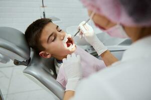 sur de soi enfant garçon séance dans dentiste chaise dans contemporain dentaire clinique tandis que méconnaissable femelle dentiste Faire médical manipulations dans le sien ouvert bouche par joue rétracteur et dentaire miroir photo