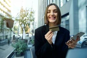 en ligne achats, éloigné travail, affaires et la finance concept. magnifique femme, entrepreneur, avec téléphone intelligent et or crédit carte , sourit à pleines dents sourire permanent contre Urbain moderne bâtiments Contexte photo