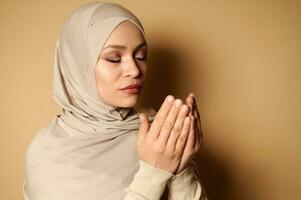 fermer portrait de une serein Jeune arabe femme dans hijab prier. religieux magnifique femme dans strict tenue prier sur beige Contexte avec copie espace photo