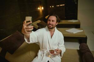 Beau Jeune européen homme dans peignoir de bain séance par jacuzzi dans bien-être spa centre, en portant tasse de à base de plantes thé et sourit à pleines dents sourire fabrication selfie ou parlant par vidéo appel tandis que relaxant dans spa recours photo