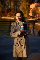 magnifique femme en portant une à emporter tasse avec une chaud boisson et une livre à la recherche à le côté tandis que en marchant dans le l'automne parc contre le Contexte de une magnifique Lac avec chute Jaune d'or feuilles photo