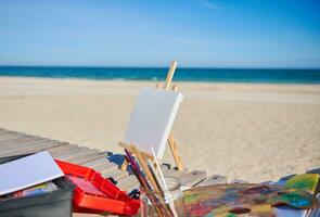 blanc Vide Toile sur une en bois table chevalet suivant à des peintures et brosses sur une en bois plate-forme chaise sur le plage contre le magnifique bleu mer Contexte photo