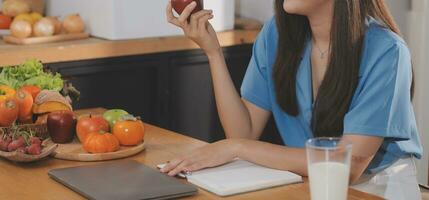 une Jeune femme avec une magnifique visage dans une bleu chemise avec longue cheveux en mangeant fruit séance à l'intérieur le cuisine à Accueil avec une portable et carnet pour relaxation, concept vacances. photo