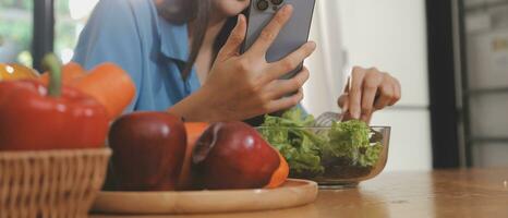 une Jeune femme avec une magnifique visage dans une bleu chemise avec longue cheveux en mangeant fruit séance à l'intérieur le cuisine à Accueil avec une portable et carnet pour relaxation, concept vacances. photo