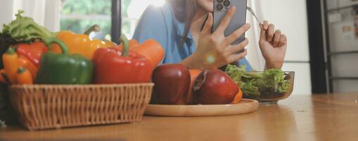 une Jeune femme avec une magnifique visage dans une bleu chemise avec longue cheveux en mangeant fruit séance à l'intérieur le cuisine à Accueil avec une portable et carnet pour relaxation, concept vacances. photo