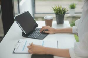 portrait d'une jeune femme asiatique pigiste à la main travaille son travail sur une tablette informatique dans un bureau moderne. faire un rapport d'analyse comptable sur les données d'investissement immobilier, le concept de systèmes financiers et fiscaux. photo