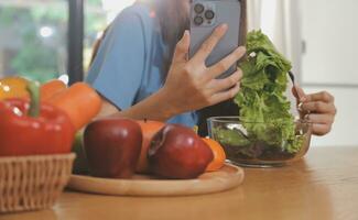 une Jeune femme avec une magnifique visage dans une bleu chemise avec longue cheveux en mangeant fruit séance à l'intérieur le cuisine à Accueil avec une portable et carnet pour relaxation, concept vacances. photo