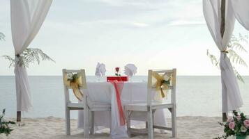 romantique dîner table ensemble et chaise préparation avec blanc thème décoration, blanc sable, clair nuageux ciel, et tranquille, pacifique, magnifique plage vue à le rivage. bien endroit pour mariage proposition photo