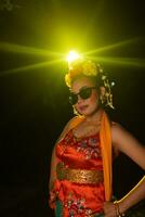une sundanais Danseur pose dans de face de une brillant lumière tandis que portant un Orange costume avec des lunettes de soleil et fleurs sur sa tête photo