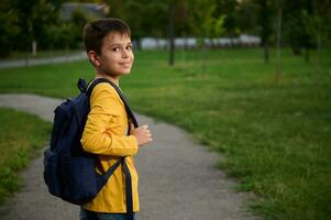 côté vue de une écolier avec cartable sac à dos en marchant sur le chemin dans Publique parc, Aller Accueil après école, mignonne souriant à le caméra photo