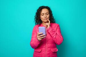 attrayant perplexe pensif aux cheveux noirs femme dans brillant encre veste met sa doigt sur lèvres et regards pensivement sur une téléphone intelligent dans sa mains, isolé sur bleu coloré Contexte avec copie espace photo