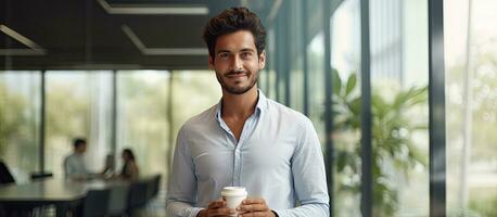 arabe homme en portant café tasse sur de soi permanent par verre mur dans Bureau et à la recherche à caméra photo