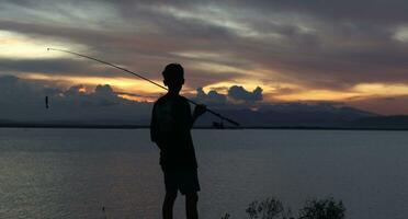 silhouette de une pêcheur avec une pêche barre dans le Lac à le coucher du soleil photo