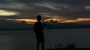 silhouette de une pêcheur avec une pêche barre dans le Lac à le coucher du soleil photo