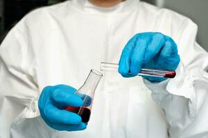 une médecin dans gants verse une rouge liquide de un tester tube dans un autre. photo