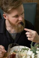 une Jeune homme avec une barbe goûts nourriture à une restaurant table photo