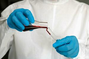 une médecin dans gants verse une rouge liquide de un tester tube dans un autre photo
