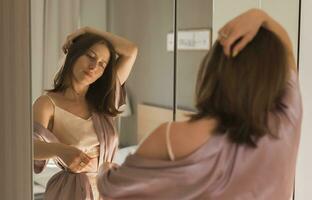 content Matin portrait. attrayant Jeune femme à la recherche dans miroir à sa appartement - réveiller en haut et Nouveau journée photo