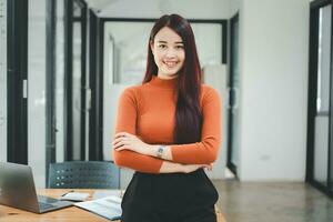 portrait de Jeune souriant femme à la recherche à caméra avec franchi bras. content fille permanent dans Créatif bureau. réussi femme d'affaires permanent dans Bureau avec copie espace. photo