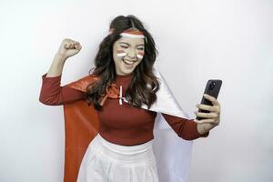 une Jeune asiatique femme avec une content réussi expression tandis que en portant sa téléphone et portant rouge haut, drapeau Bandeau et cap isolé par blanc Contexte. l'indonésie indépendance journée concept. photo